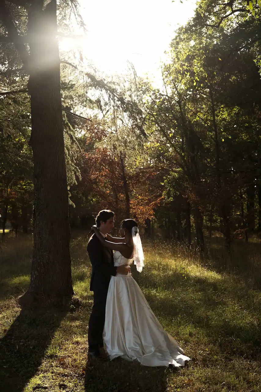 Un couple de marié·es s'enlace tendrement dans une forêt baignée par la lumière dorée du soleil couchant. La mariée porte une robe blanche avec un voile, tandis que le marié est vêtu d'un costume noir.