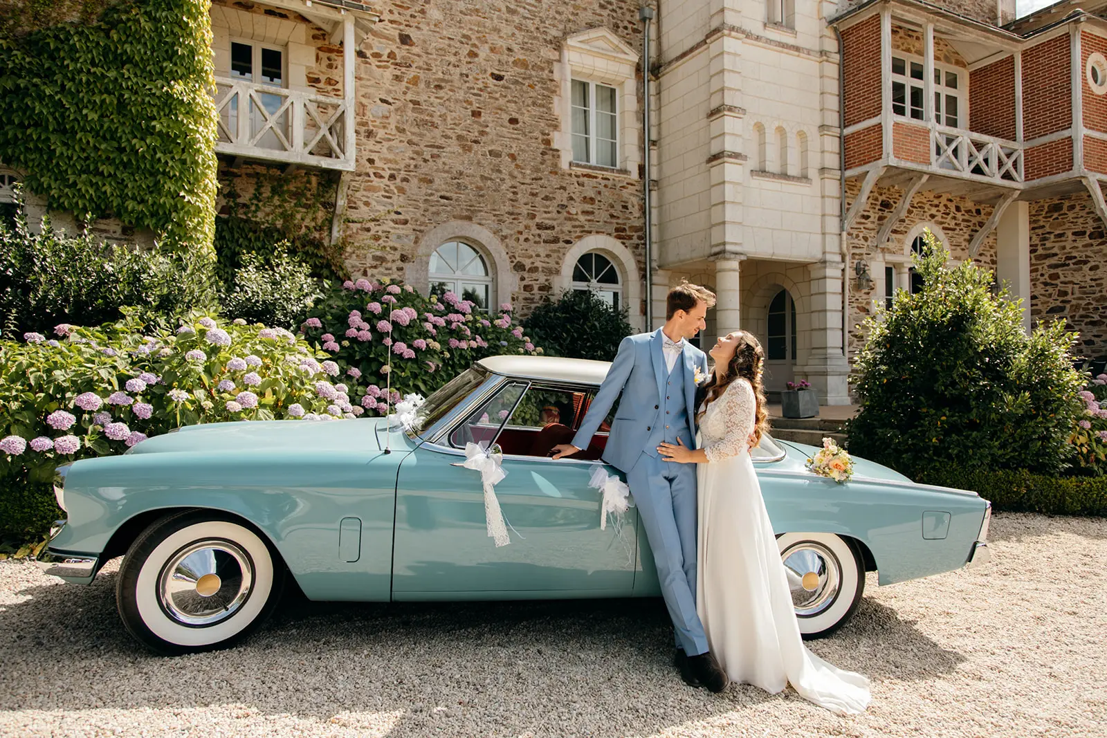 Un couple de mariés se regarde tendrement devant une voiture de collection bleu ciel, garée sur une allée gravillonnée devant un château en pierre. La mariée porte une robe blanche en dentelle et le marié un costume bleu clair assorti à la voiture décorée de nœuds blancs.