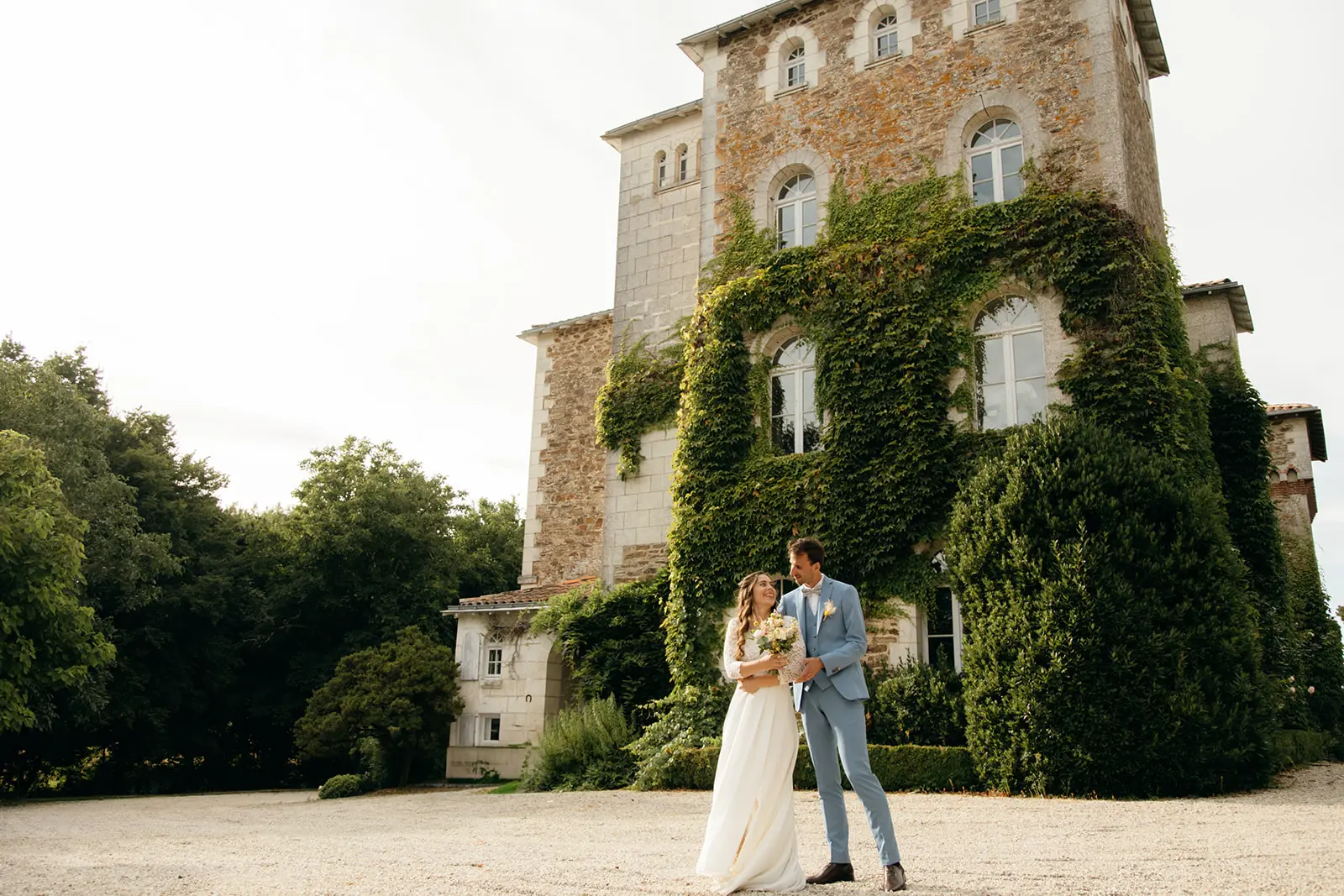 Un couple de mariés se regarde avec tendresse devant un château en pierre recouvert de vigne vierge. La mariée porte une robe blanche et le marié un costume bleu clair.