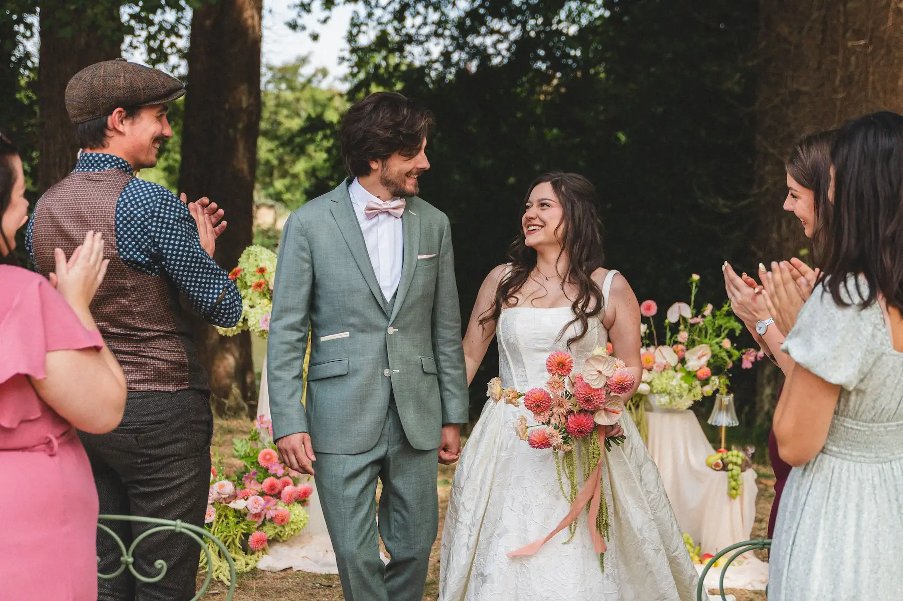 Un couple de jeunes mariés se tient la main et se regarde avec tendresse en marchant, entouré d'invités qui les applaudissent à la fin de leur cérémonie en extérieur. La mariée porte une robe blanche et tient un bouquet de fleurs roses et verts, tandis que le marié est vêtu d'un costume vert clair.