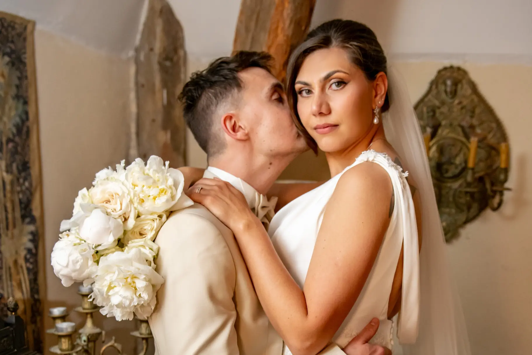 Une mariée en robe blanche tenant un bouquet de pivoines regarde l'objectif tandis que son époux, en costume beige, l'embrasse tendrement sur la joue.