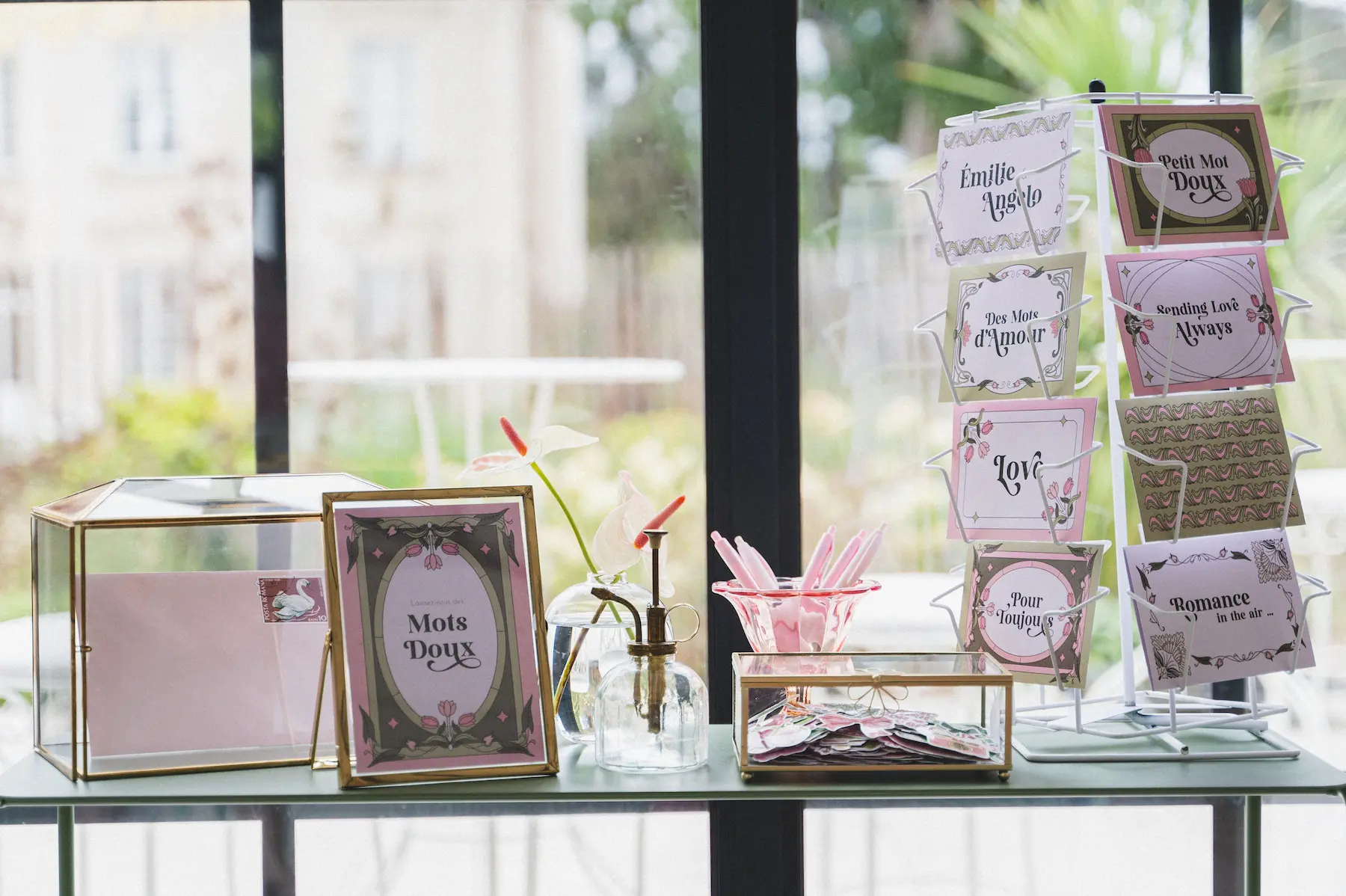 Un bar à cartes postales pour un mariage, composé d'une urne en verre, de stylos, de cartes décorées et d'un présentoir avec des messages tels que « Mots Doux », « Pour Toujours » et « Romance in the air ».